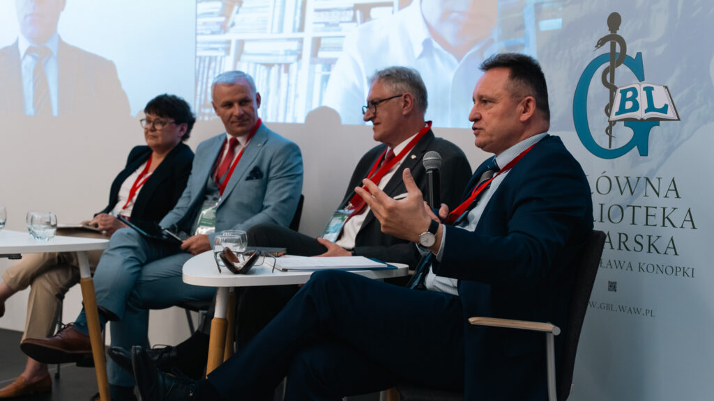 Panel dyskusyjny podczas międzynarodowej konferencji medycznej. Kilku prelegentów siedzi przy stolikach, jeden z nich wypowiada się z mikrofonem, a w tle widoczna jest projekcja z uczestnikami online oraz logo Głównej Biblioteki Lekarskiej.