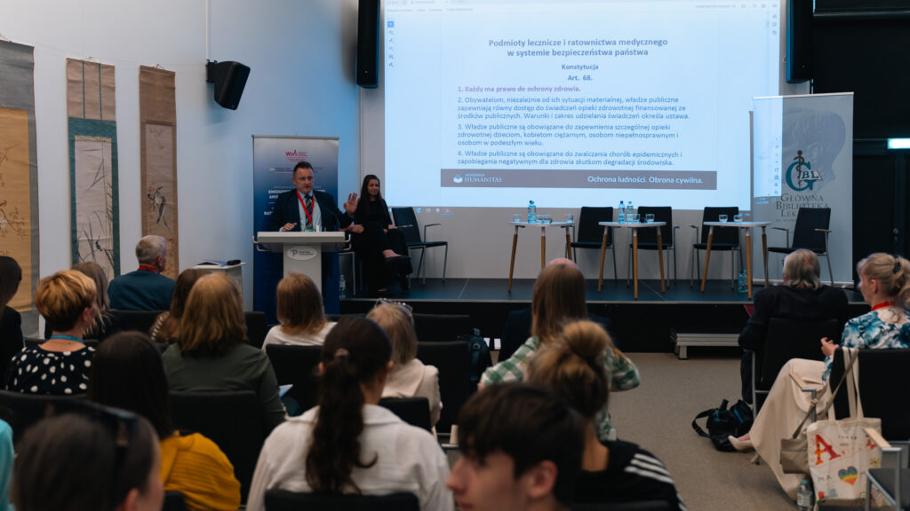 Prelegent prowadzi wystąpienie podczas międzynarodowej konferencji dotyczącej medycyny, edukacji i ratownictwa medycznego. Na dużym ekranie widoczny jest slajd o podmiotach leczniczych i ratownictwie medycznym w systemie bezpieczeństwa państwa, a publiczność siedzi w sali konferencyjnej.