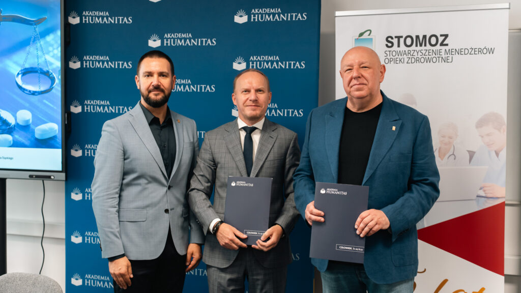 Trzej mężczyźni stoją z teczkami z porozumieniem na tle ścianki Akademii Humanitas oraz roll-upu STOMOZ.