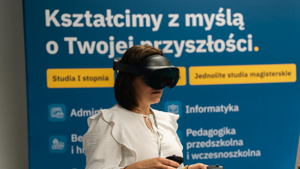 Kobieta w okularach VR stoi na tle ścianki Akademii Humanitas z napisem "Kształcimy z myślą o Twojej przyszłości".