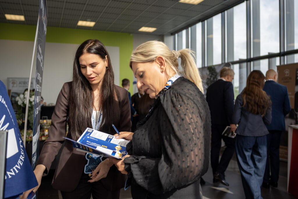 Przedstawicielka Akademii Humanitas rozmawia z uczestniczką Prime HR Forum przy stoisku uczelni. Kobiety oglądają materiały promocyjne, a w tle widoczni są inni uczestnicy wydarzenia oraz stoiska wystawiennicze.