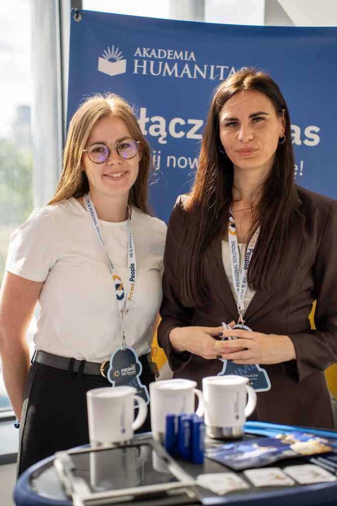 Dwie przedstawicielki pozują przy stoisku Akademii Humanitas podczas Prime HR Forum. Na pierwszym planie widoczne są kubki, długopisy i materiały promocyjne uczelni, a w tle znajduje się baner Akademii Humanitas z hasłem „Dołącz do nas i zacznij nową przygodę!”.
