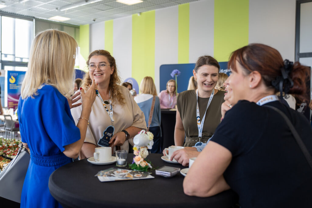 Uczestniczki Prime HR Forum rozmawiają przy stoliku networkingowym podczas przerwy w wydarzeniu. Kobiety mają identyfikatory konferencyjne, trzymają filiżanki z kawą i uśmiechają się, a w tle widoczne są inne osoby oraz stoiska wystawiennicze.
