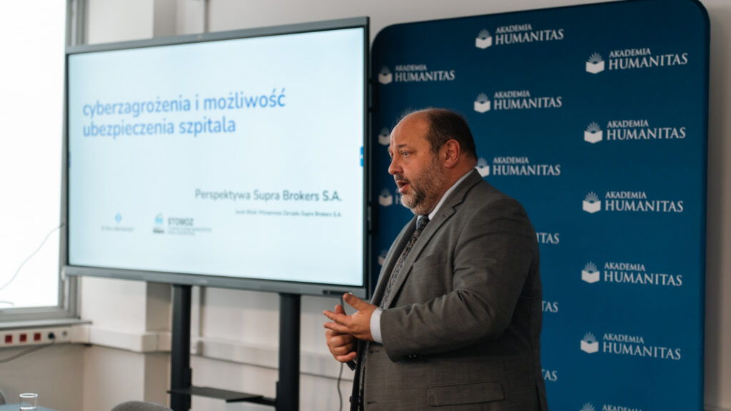 Prelegent stoi na tle ścianki Akademii Humanitas. W tle telewizor z widocznym tematem wystąpienia "cyberzagrożenia i możliwość ubezpieczenia szpitala".