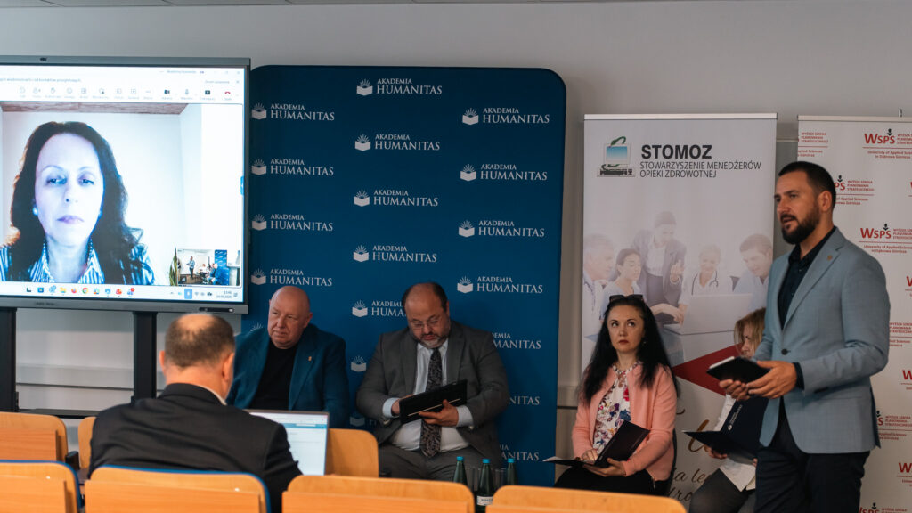 Panel konferencyjny Akademii Humanitas z udziałem prelegentów stacjonarnych i zdalnych. Jeden z uczestników przemawia stojąc z notesem, pozostali siedzą przy stole, a na dużym ekranie widoczna jest osoba łącząca się online.