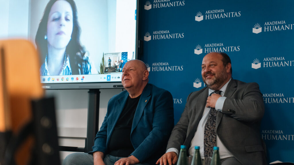 Dwóch panelistów siedzi na tle ścianki Akademii Humanitas podczas wydarzenia konferencyjnego. Po lewej stronie na dużym ekranie widoczna jest prelegentka łącząca się online, a na pierwszym planie widać butelki z wodą i fragment wyposażenia sali.