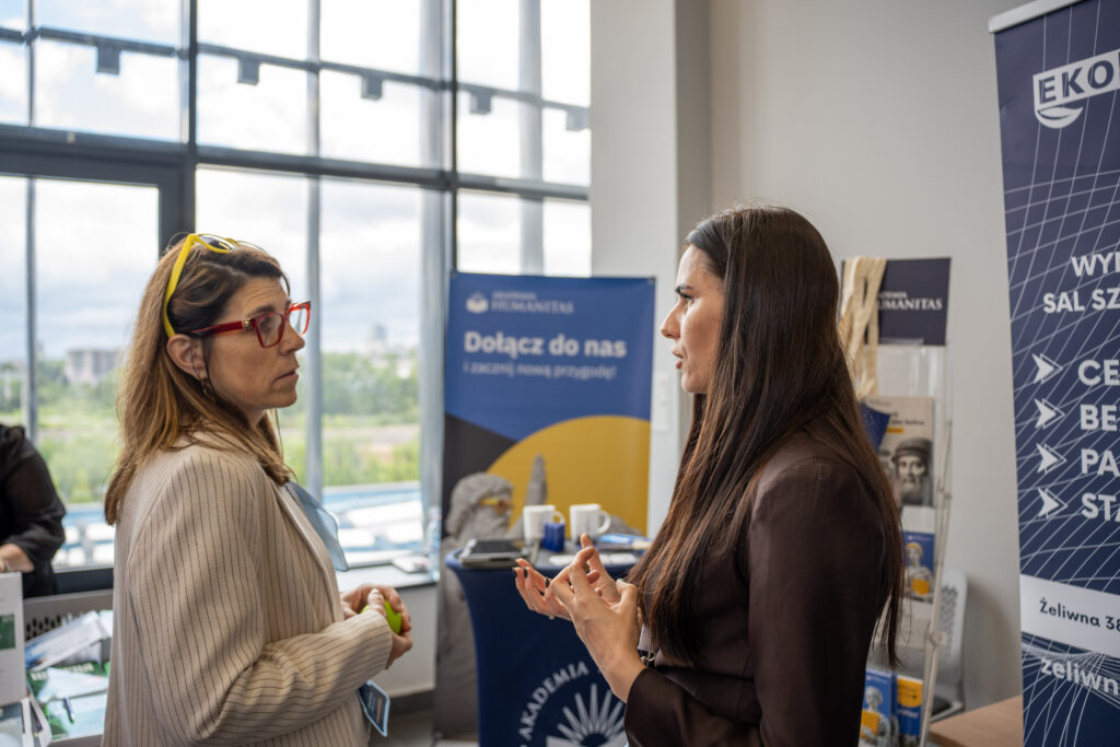 Uczestniczki Prime HR Forum rozmawiają przy stoliku networkingowym podczas przerwy w wydarzeniu. Kobiety mają identyfikatory konferencyjne, trzymają filiżanki z kawą i uśmiechają się, a w tle widoczne są inne osoby oraz stoiska wystawiennicze.