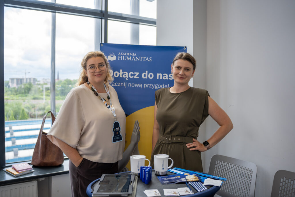 Dwie kobiety pozują przy stoisku Akademii Humanitas podczas Prime HR Forum. Na stoliku widoczne są materiały promocyjne, kubki i gadżety uczelni, a w tle znajduje się baner z hasłem „Dołącz do nas i zacznij nową przygodę!