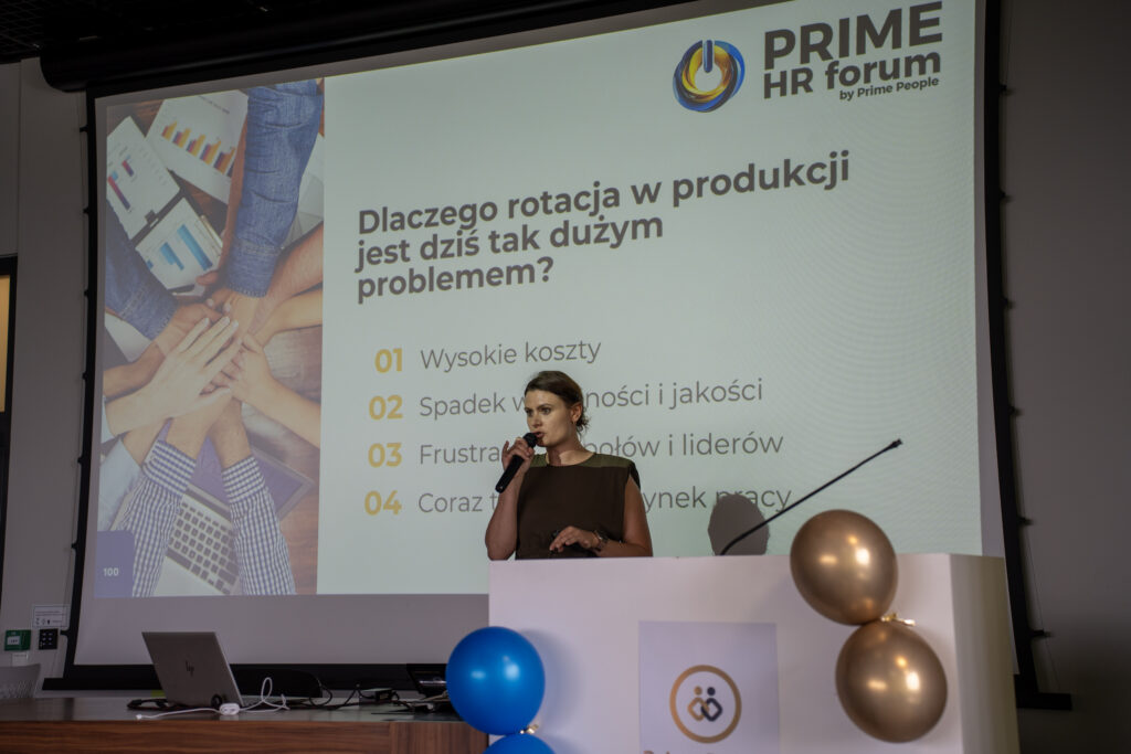 Prelegentka podczas Prime HR Forum omawia problem rotacji pracowników w produkcji, wskazując na slajd z pytaniem „Dlaczego rotacja w produkcji jest dziś tak dużym problemem?”. Publiczność siedzi w sali konferencyjnej, a na ekranie widoczne są punkty dotyczące wysokich kosztów, spadku wydajności i jakości oraz frustracji zespołów.