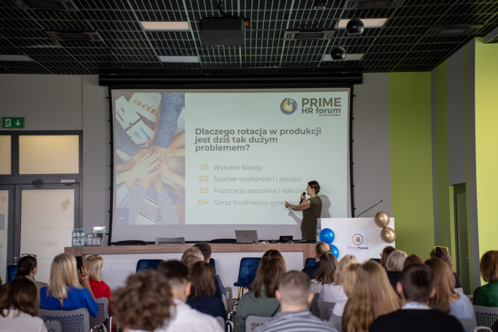 Prelegentka podczas Prime HR Forum omawia problem rotacji pracowników w produkcji, wskazując na slajd z pytaniem „Dlaczego rotacja w produkcji jest dziś tak dużym problemem?”. Publiczność siedzi w sali konferencyjnej, a na ekranie widoczne są punkty dotyczące wysokich kosztów, spadku wydajności i jakości oraz frustracji zespołów.