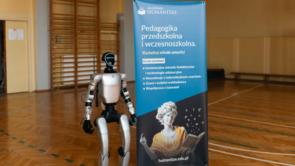 Humanoidalny robot stoi obok banera Akademii Humanitas promującego kierunek „Pedagogika przedszkolna i wczesnoszkolna”. Zdjęcie wykonano w sali gimnastycznej, w której widoczna jest drewniana podłoga, drabinki i linie boiska.