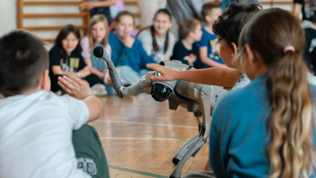 Dzieci siedzą na podłodze sali gimnastycznej i obserwują robota kroczącego z bliska. Kilkoro dzieci wyciąga ręce w stronę robota i dotyka jego obudowy podczas pokazu technologicznego.