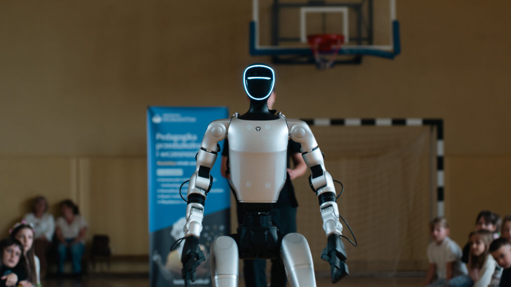 Humanoidalny robot stoi na środku sali gimnastycznej podczas pokazu dla dzieci. W tle widoczne są uczniowie siedzący po bokach, bramka sportowa, kosz do koszykówki oraz baner Akademii Humanitas.