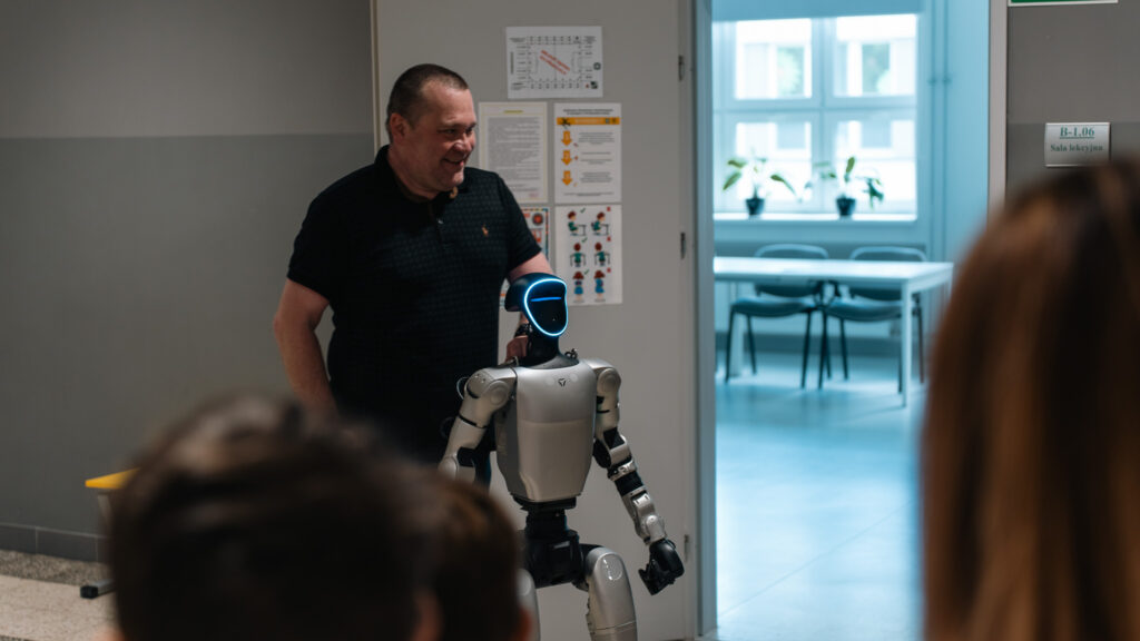 Prowadzący prezentuje humanoidalnego robota. Robot z podświetlaną twarzą stoi przed uczestnikami, a w tle widać wejście do sali lekcyjnej i uśmiechniętego prowadzącego.