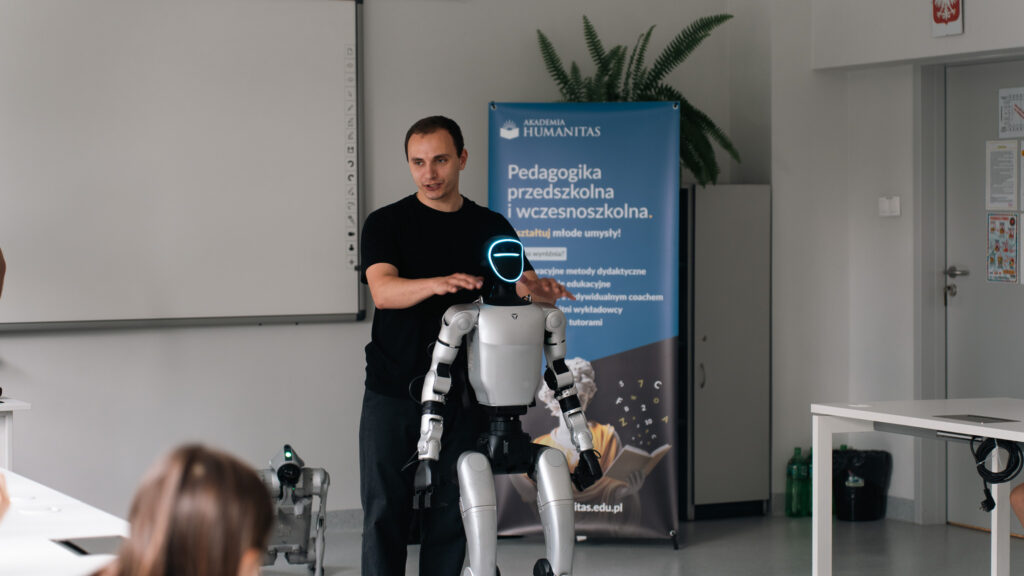 Prowadzący prezentuje humanoidalnego robota. Robot stoi przed uczestnikami, a w tle widoczny jest baner kierunku „Pedagogika przedszkolna i wczesnoszkolna” oraz drugi robot kroczący.