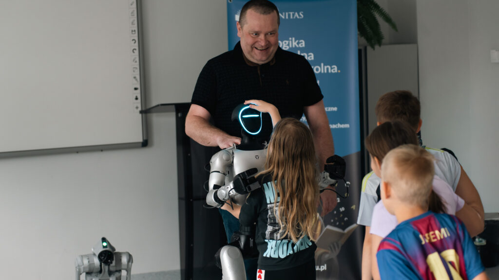 Dzieci z bliska oglądają humanoidalnego robota podczas pokazu. Jedno z dzieci dotyka podświetlanej głowy robota, obok stoi uśmiechnięty prowadzący, a na podłodze widoczny jest także robot kroczący.