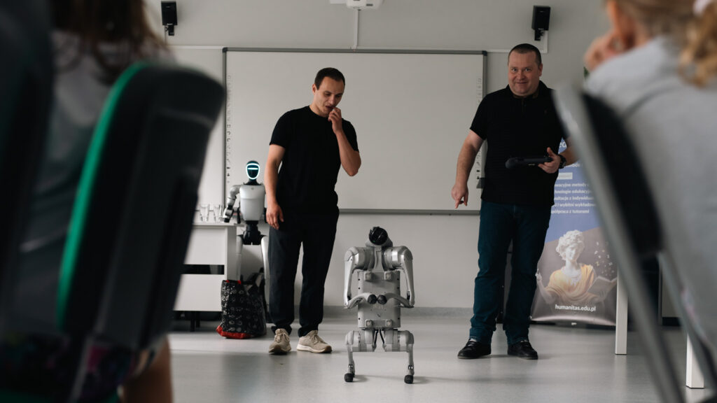 Pokaz robotów humanoidalnych. Dwóch prowadzących prezentuje urządzenia przed uczestnikami, a na pierwszym planie widoczny jest robot kroczący stojący na środku sali.