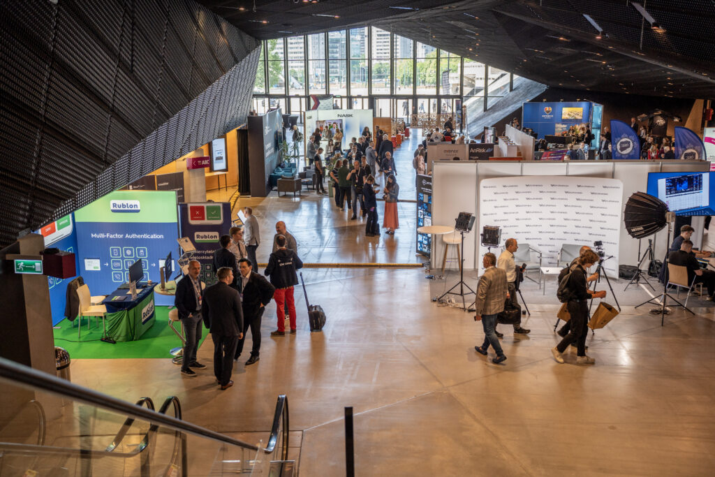 Widok z góry na przestrzeń wystawienniczą podczas Cybersec Expo & Forum. Uczestnicy rozmawiają przy stoiskach firm i instytucji związanych z cyberbezpieczeństwem, a w sali widoczne są banery, ekrany prezentacyjne oraz strefy networkingowe.