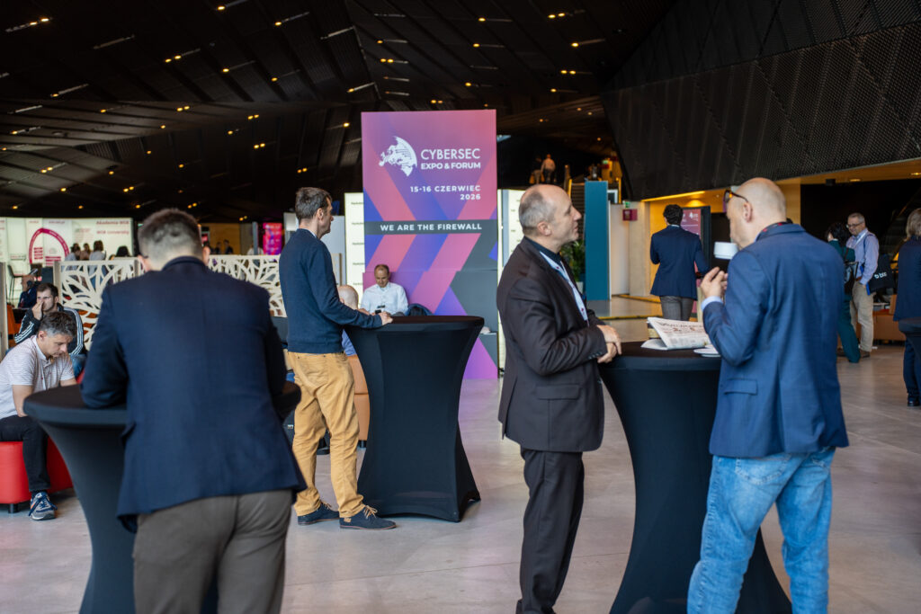 Uczestnicy Cybersec Expo & Forum rozmawiają podczas przerwy networkingowej w przestrzeni konferencyjnej. W centrum widoczny jest wysoki baner wydarzenia z hasłem „We are the firewall”, a wokół niego stoją osoby przy stolikach koktajlowych.