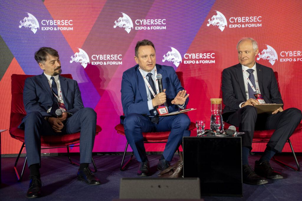 Panel dyskusyjny podczas Cybersec Expo & Forum. Trzech prelegentów siedzi na czerwonych fotelach, jeden z nich mówi do mikrofonu i gestykuluje, a w tle widoczny jest ekran z logotypami wydarzenia.