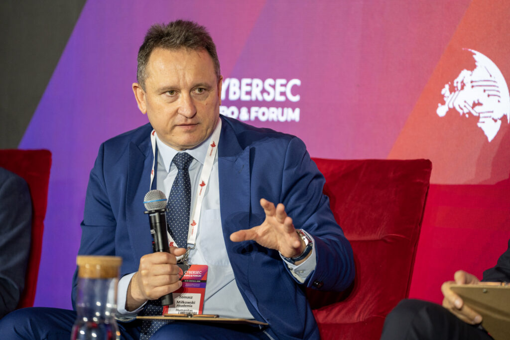 Prelegent w niebieskim garniturze wypowiada się do mikrofonu podczas panelu na Cybersec Expo & Forum. Siedzi na czerwonym fotelu, gestykuluje dłonią, a w tle widoczny jest ekran z logotypem wydarzenia.