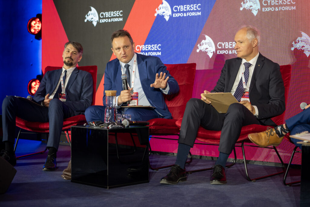 Panel dyskusyjny podczas Cybersec Expo & Forum. Trzech prelegentów siedzi na czerwonych fotelach, jeden z nich mówi do mikrofonu i gestykuluje, a w tle widoczny jest ekran z logotypami wydarzenia.