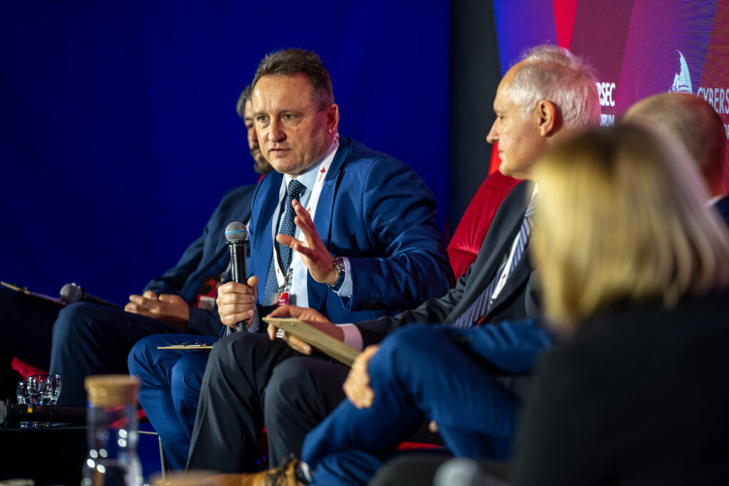 Panel dyskusyjny podczas konferencji Cyber Science Expo. Jeden z prelegentów w niebieskim garniturze wypowiada się do mikrofonu, a obok siedzą pozostali uczestnicy panelu na czerwonych fotelach.