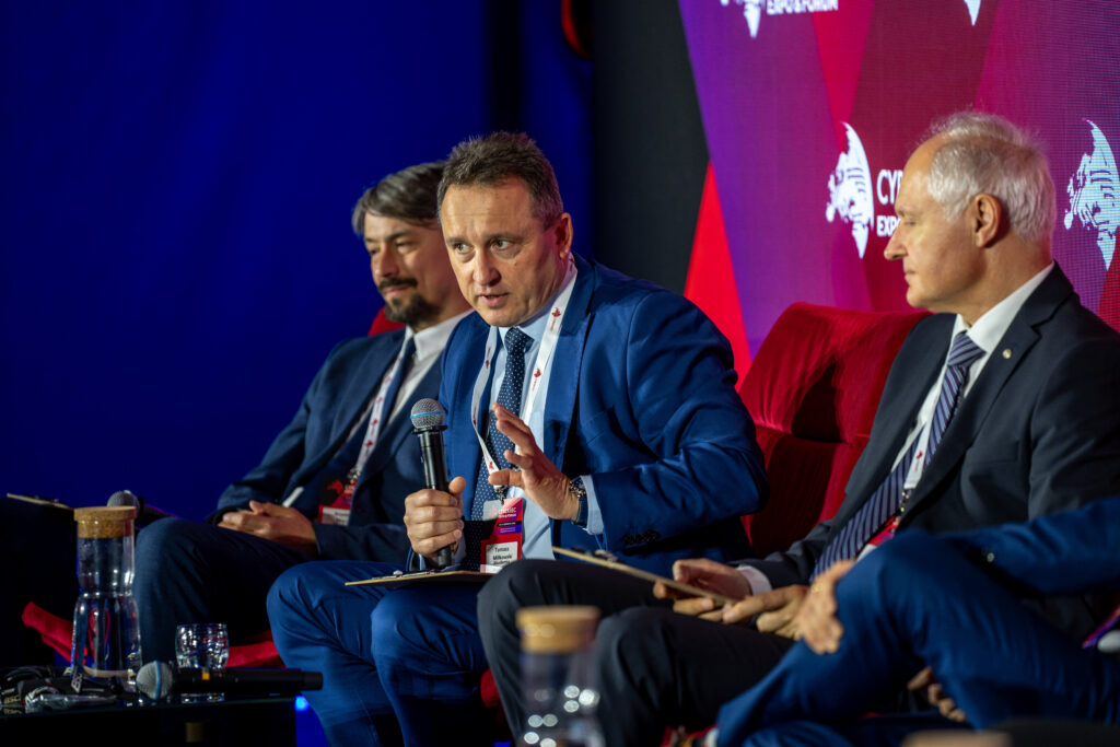 Panel dyskusyjny podczas konferencji Cyber Science Expo. Jeden z prelegentów w niebieskim garniturze wypowiada się do mikrofonu, a obok siedzą pozostali uczestnicy panelu na czerwonych fotelach.