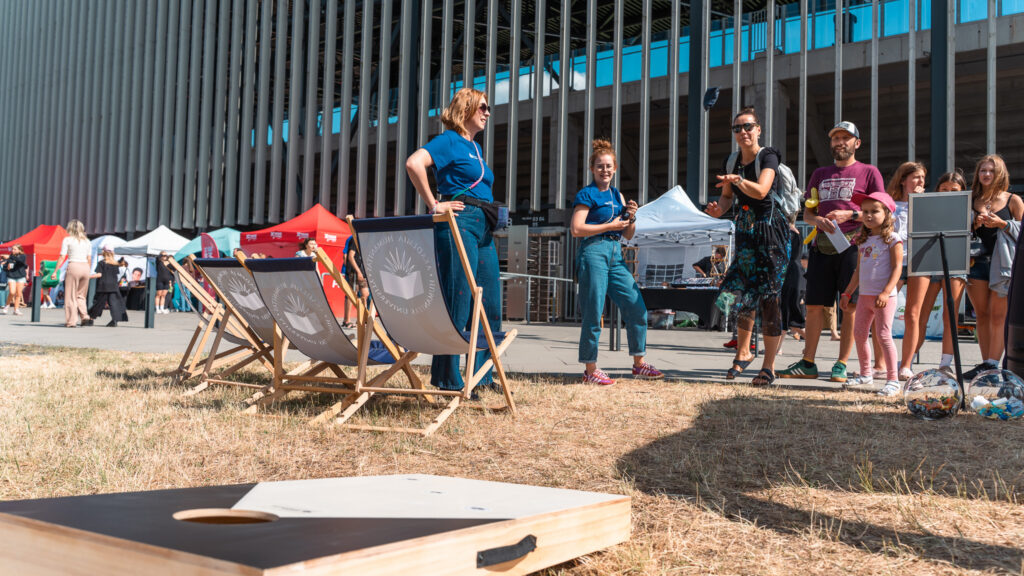 Kobieta rzuca woreczkiem do celu podczas gry w cornhole. Za nią stoją dzieci i mężczyzna i czekają na swoją kolej. 