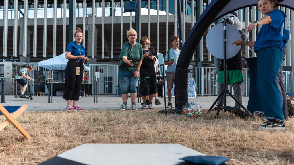 3 osoby stoją w kolejce do gry cornhole. Po lewej stronie stoi kobieta i podaje woreczki. Po prawej Kobieta wskazuje palcem na grę. 