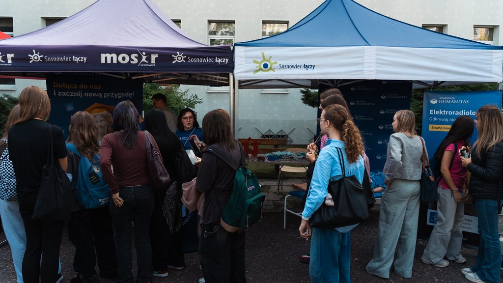 Stoisko Akademii Humanitas podczas plenerowego wydarzenia profilaktyczno-edukacyjnego. Grupa młodzieży odwiedza stanowiska pod namiotami Miasta Sosnowiec i Akademii Humanitas, gdzie przedstawicielki uczelni rozmawiają z uczestniczkami i przekazują materiały informacyjne.