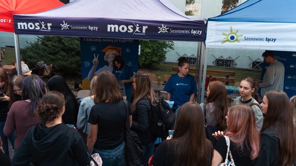 Stoisko Akademii Humanitas podczas plenerowego wydarzenia profilaktyczno-edukacyjnego. Grupa młodzieży odwiedza stanowiska pod namiotami Miasta Sosnowiec i Akademii Humanitas, gdzie przedstawicielki uczelni rozmawiają z uczestniczkami i przekazują materiały informacyjne.