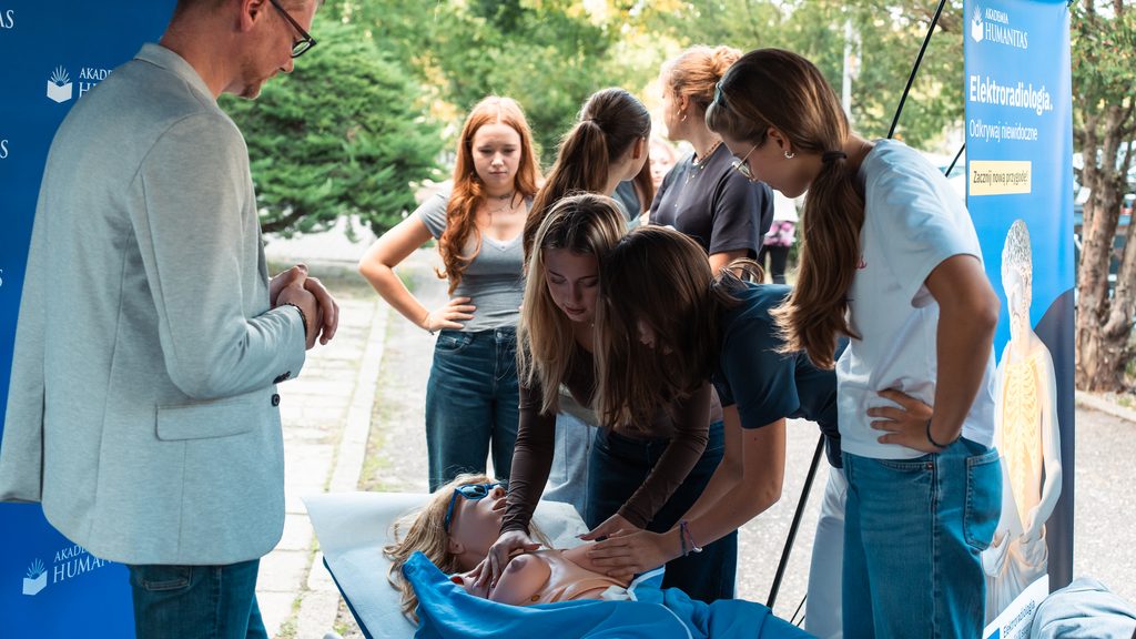 Uczennice ćwiczą na fantomie podczas plenerowego wydarzenia edukacyjno-profilaktycznego Akademii Humanitas. Obok stoi prowadzący, a w tle widoczne są banery uczelni, w tym grafika promująca kierunek elektroradiologia.