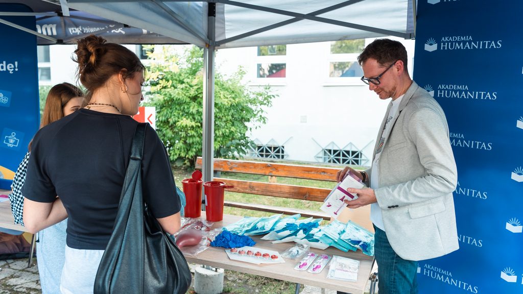 Przedstawiciel Akademii Humanitas omawia materiały edukacyjne przy stoisku profilaktycznym. Na stole widoczne są modele anatomiczne, rękawiczki, pakiety medyczne i ulotki, a uczestniczki wydarzenia zapoznają się z wyposażeniem stoiska.