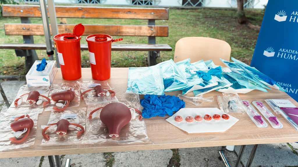 Materiały edukacyjne i modele anatomiczne rozłożone na stole stoiska Akademii Humanitas podczas wydarzenia profilaktycznego. Widoczne są m.in. modele narządów rodnych, rękawiczki, pakiety medyczne, pojemniki na odpady oraz akcesoria wykorzystywane do działań edukacyjnych z zakresu profilaktyki zdrowia kobiet.