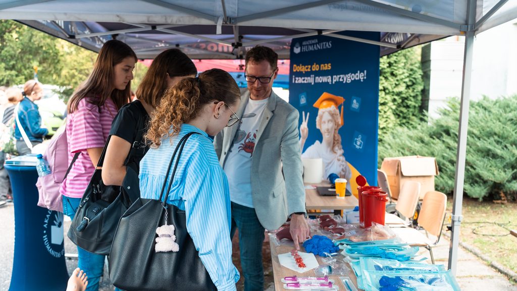 Stoisko Akademii Humanitas podczas wydarzenia plenerowego poświęconego profilaktyce zdrowotnej. Uczestniczki rozmawiają z przedstawicielem uczelni i oglądają materiały edukacyjne oraz drobne akcesoria na stole, a w tle widoczny jest baner promocyjny Akademii Humanitas.