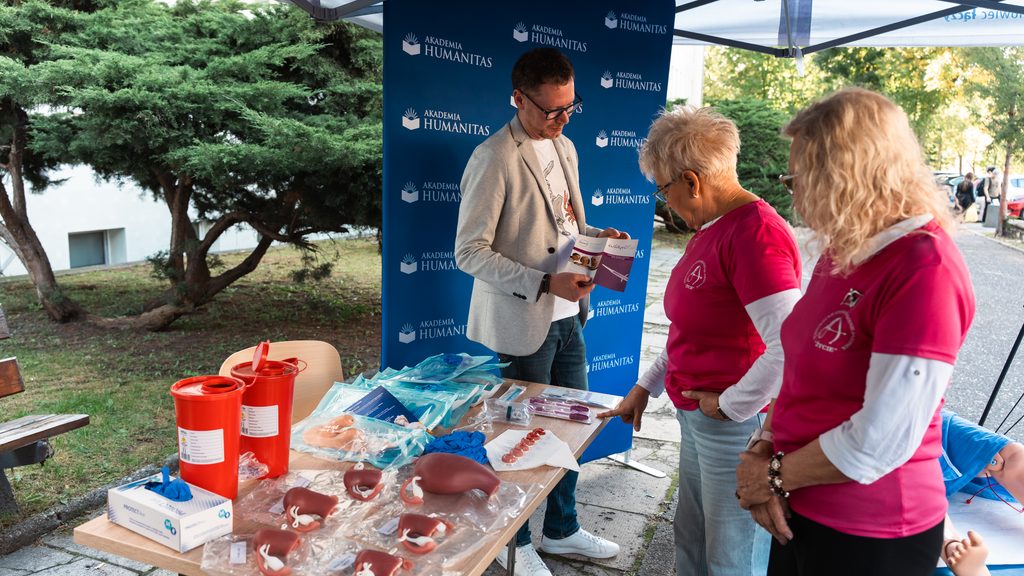 Przedstawiciel Akademii Humanitas omawia materiały edukacyjne przy stoisku profilaktycznym podczas wydarzenia plenerowego. Na stole widoczne są modele anatomiczne, pakiety medyczne i ulotki, a obok stoją uczestniczki wydarzenia w różowych koszulkach.