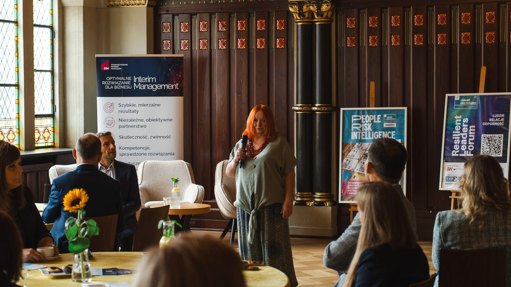 Prelegentka z mikrofonem zabiera głos podczas spotkania w zabytkowej sali. Uczestnicy siedzą przy okrągłych stołach, a w tle znajdują się materiały promocyjne wydarzenia.
