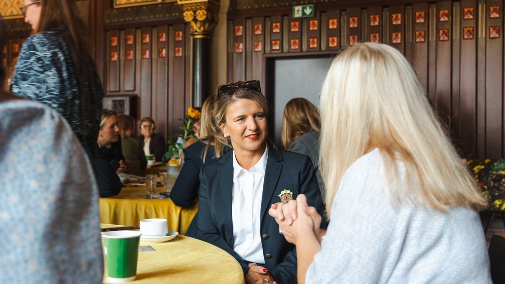 Uczestniczki wydarzenia rozmawiają przy okrągłym stole w zabytkowej sali. Spotkanie odbywa się w atmosferze networkingu i wymiany doświadczeń.