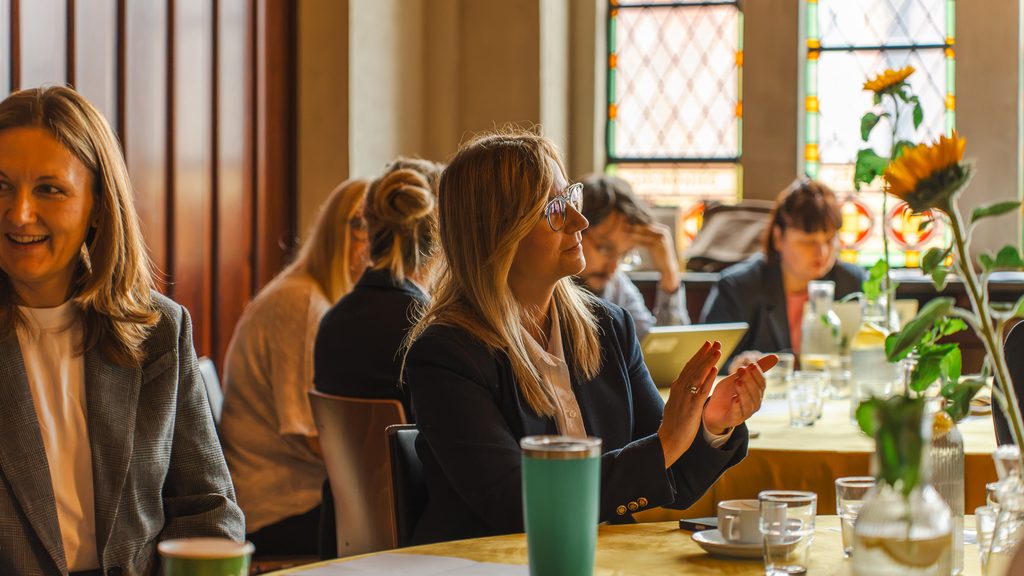 Uczestniczki Resilient Leaders Forum słuchają wystąpienia przy okrągłym stole. Wnętrze sali podkreślają witraże, drewniane wykończenia i dekoracje ze słonecznikami.