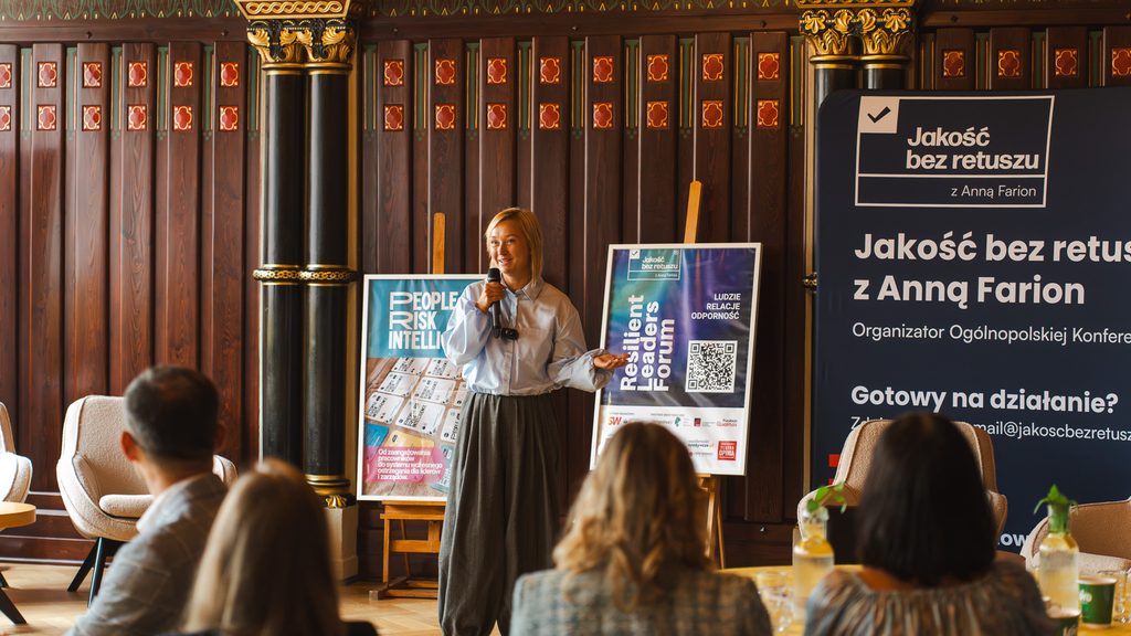 Prelegentka prowadzi wystąpienie podczas Resilient Leaders Forum w eleganckiej, zabytkowej sali. Stoi z mikrofonem przed publicznością, a w tle widoczne są plakaty wydarzenia oraz duży baner „Jakość bez retuszu”.
