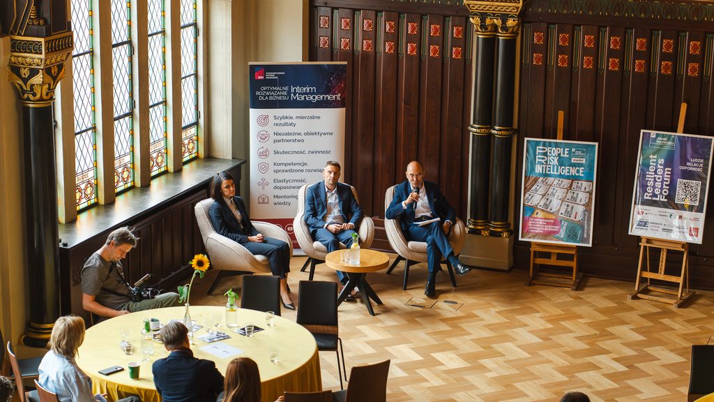 Panel dyskusyjny podczas Resilient Leaders Forum w zabytkowej sali konferencyjnej. Prelegenci siedzą na fotelach przed publicznością, a w tle widoczne są banery partnerów i plakaty wydarzenia.