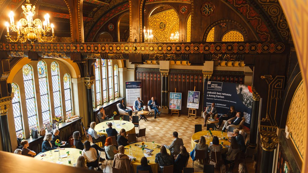 Uczestnicy Resilient Leaders Forum biorą udział w spotkaniu w eleganckiej, historycznej sali z bogato zdobionym wnętrzem. Wydarzenie odbywa się przy okrągłych stołach, a dyskusja prowadzona jest w formule panelowej.