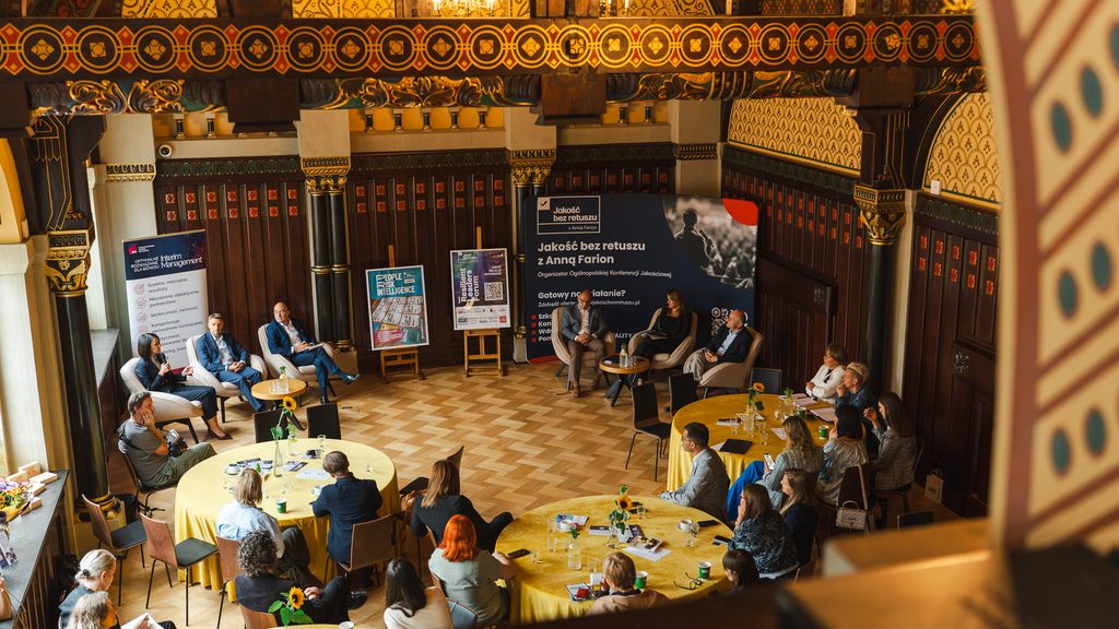 Widok z góry na panel dyskusyjny Resilient Leaders Forum w zabytkowej sali. Uczestnicy siedzą przy okrągłych stołach, a prelegenci prowadzą rozmowę na scenie otoczonej materiałami wydarzenia.