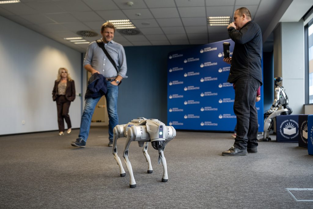 Robotyczny pies prezentowany podczas wydarzenia Akademii Humanitas. W tle widoczni są uczestnicy spotkania, fotograf oraz ścianka z logotypami uczelni.