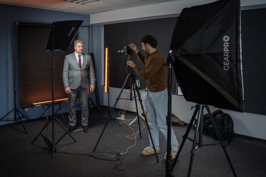 Operator fotografuje uczestnika wydarzenia w profesjonalnym studiu zdjęciowym. W pomieszczeniu widoczne są lampy, tła fotograficzne oraz aparat ustawiony na statywie.