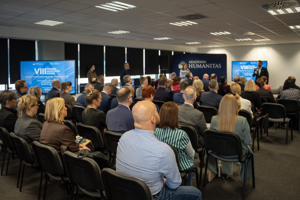 Publiczność uczestniczy w wydarzeniu Akademii Humanitas. W sali konferencyjnej widoczne są rzędy słuchaczy, ekrany z grafiką VIII Kongresu Praktyków Biznesu oraz prowadzący stojący przed uczestnikami.