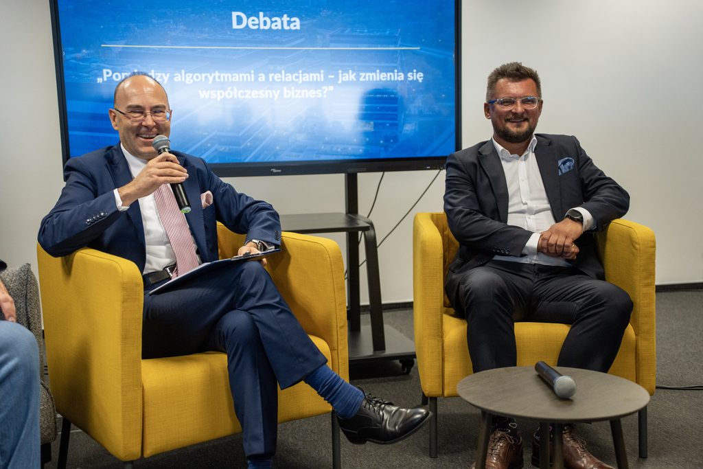 Dwóch uczestników debaty siedzi w żółtych fotelach podczas wydarzenia Akademii Humanitas. Jeden z nich mówi do mikrofonu, a w tle widoczny jest ekran z tematem debaty „Pomiędzy algorytmami a relacjami – jak zmienia się współczesny biznes?”.