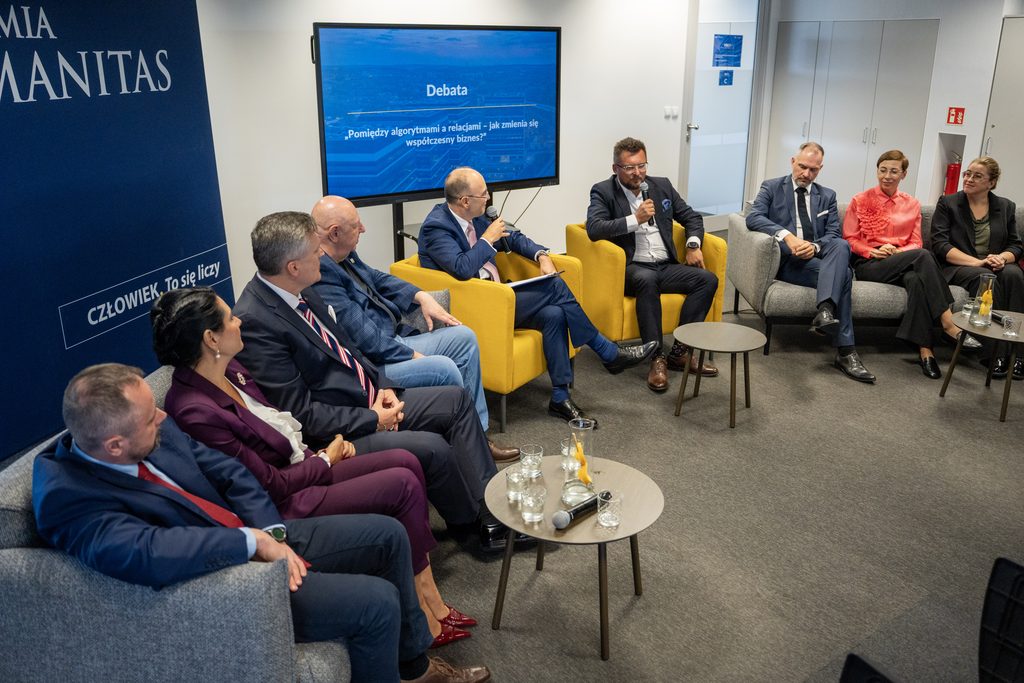 Paneliści biorą udział w debacie biznesowej organizowanej przez Akademię Humanitas. Uczestnicy siedzą w półokręgu na fotelach i kanapach, a jeden z prelegentów wypowiada się do mikrofonu.