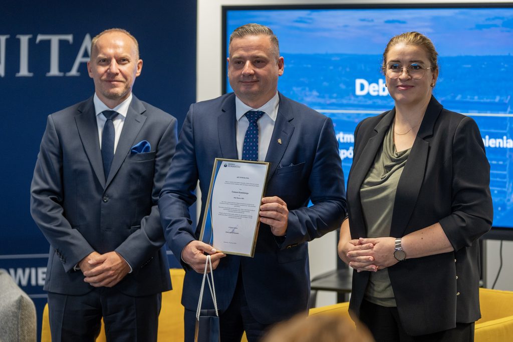 Przedstawiciele Akademii Humanitas pozują z uczestnikiem wydarzenia, który trzyma pamiątkowy akt powołania i torbę upominkową. W tle widoczny jest ekran z grafiką debaty oraz elementy identyfikacji wizualnej uczelni.